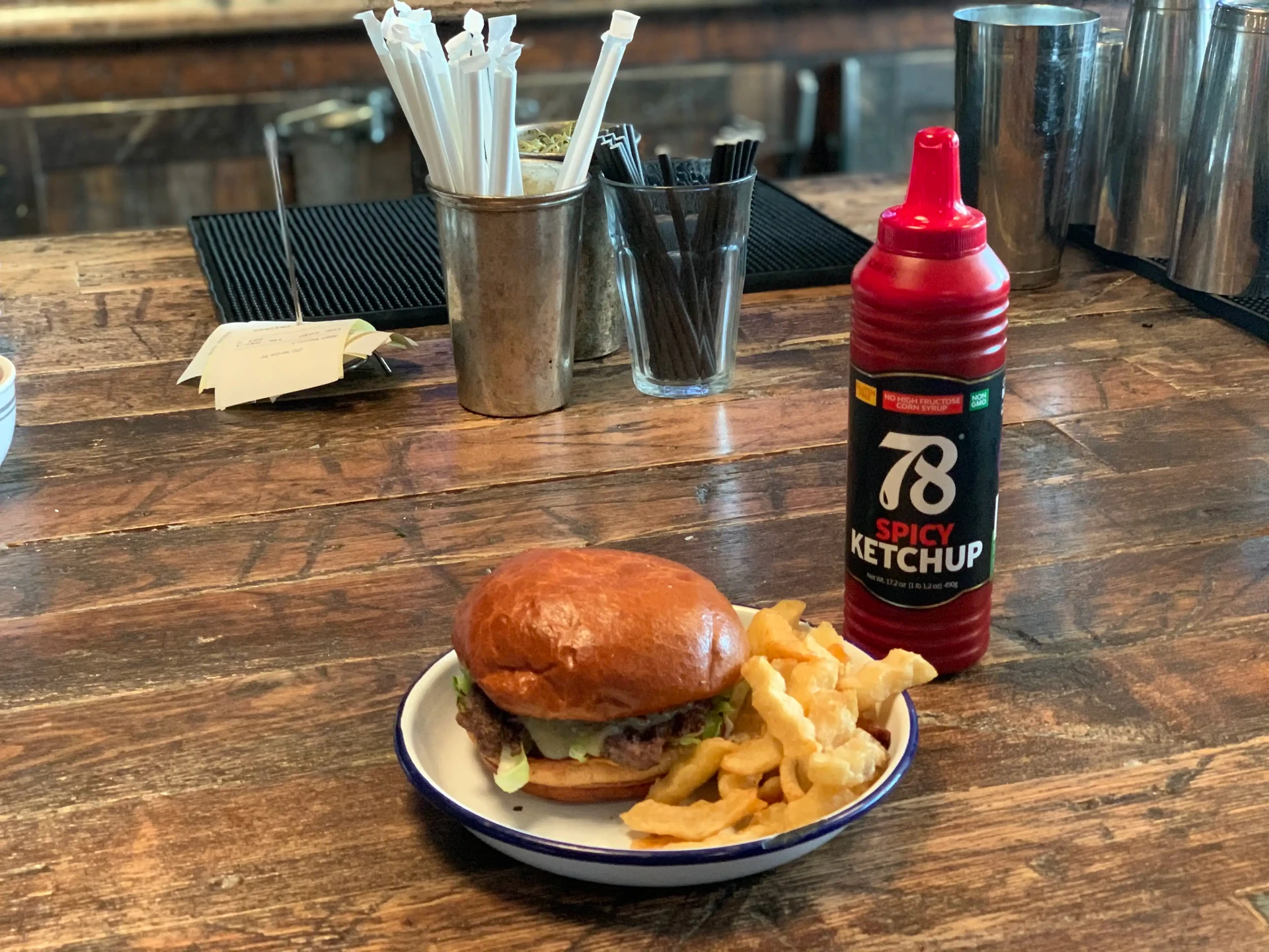 78 Brand Spicy Ketchup bottle on a wooden bar top next to a burger and crinkle cut fries during foodservice trial.